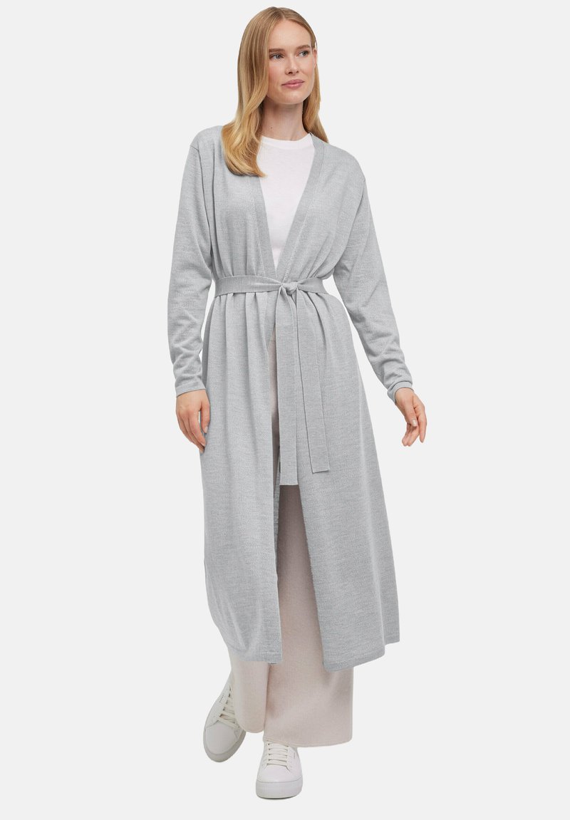 Jasnoszary długi kardigan z długimi rękawami, otwarty z przodu, z paskiem w talii. Miękka, teksturowana tkanina, nałożona na białą bluzkę i jasną spódnicę.