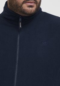 Chaqueta de forro polar azul marino con cuello alto, cremallera completa y un pequeño logo en el lado izquierdo. Textura suave y diseño cálido, adecuado para superponer.