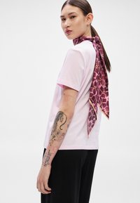 Ροζ κοντομάνικη μπλούζα συνδυασμένη με μωβ κασκόλ με σχέδια, δεμένο στον λαιμό. Μαύρο παντελόνι και ορατός βραχίονας με τατουάζ.