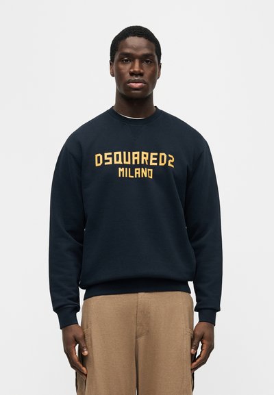 Άνδρας που φοράει μπλε φούτερ με κίτρινο κείμενο "DSQUARED2 MILANO" και μπεζ παντελόνι σε απλό ανοιχτό φόντο.