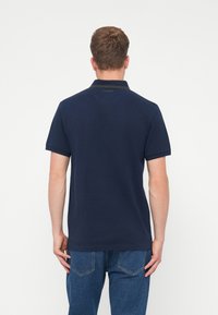 Tengerészkék póló ing sima pamut anyagból, klasszikus gallérral és bordás mandzsettával, kontrasztos zöld díszítéssel.