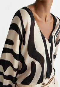 Pullover a V in beige con motivi a onda neri. Tessuto leggero lavorato a maglia con una consistenza leggermente trasparente. Maniche ampie e vestibilità rilassata.