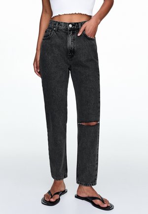 Vrouw draagt zwarte high-waist spijkerbroek met scheur op de rechterknie, gecombineerd met een wit cropped top en zwarte teenslippers.