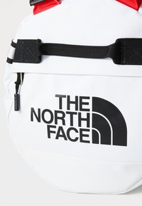White bag with black logo "THE NORTH FACE," featuring red straps and black accents. Made from durable materials with a textured finish.