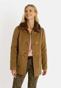 Abrigo de lana marrón con un cuello de piel sintética, cierres de tipo toggle en color beige y dos bolsillos frontales, combinado con un suéter rosa y pantalones de estampado de leopardo.