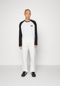 Camicia a maniche lunghe con maniche raglan nere e corpo bianco, con logo sul petto. Pantaloni bianchi e sneakers nere completano il look.