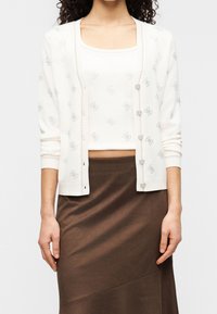 Femme portant un cardigan blanc à motifs et un top assorti avec des boutons argentés, associé à une jupe marron taille haute sur un fond uni.