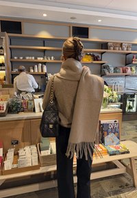 Femme portant un châle beige et un sac matelassé noir, debout au comptoir d'un café avec un barista et des étagères remplies de produits emballés et de livres.
