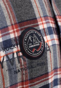 Geruite stof met rode, blauwe, grijze en witte lijnen, voorzien van een ronde patch met de tekst "Rocky Mountains in the middle of nowhere, CM Vancouver."