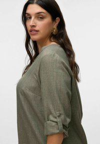Blouse en lin texturé vert olive, à manches trois quarts, avec une coupe ample et un détail de poche subtil du côté gauche.