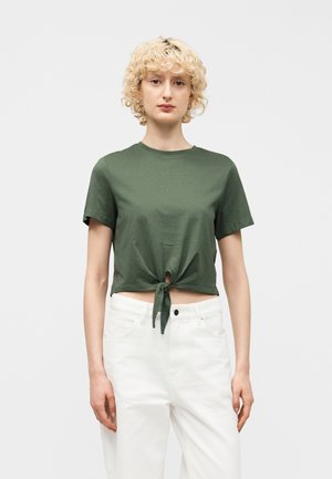 Donna con capelli biondi ricci indossa un top corto a maniche corte verde oliva annodato e pantaloni bianchi a vita alta, in piedi davanti a uno sfondo semplice.