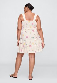 Vestido creme com padrões coloridos de conchas e sol, apresentando alças largas e uma bainha em camadas, feito de tecido leve e texturizado.