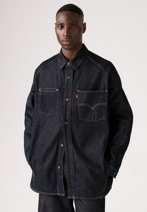 Dark denim shirt with a button-up front, pointed collar, and two chest pockets. Features yellow stitching and a relaxed fit.