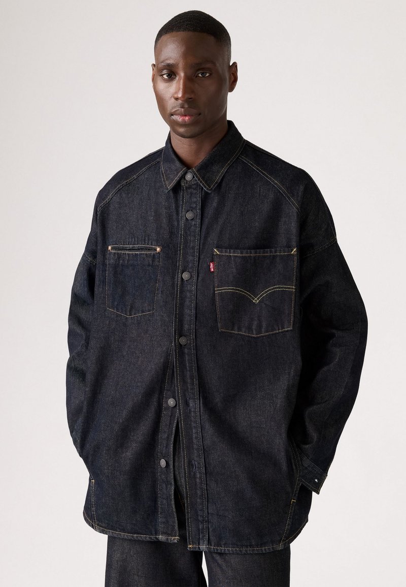 Donkerdenim shirt met een knoopsluiting aan de voorkant, puntige kraag en twee borstzakken. Heeft gele stiksels en een relaxte pasvorm.
