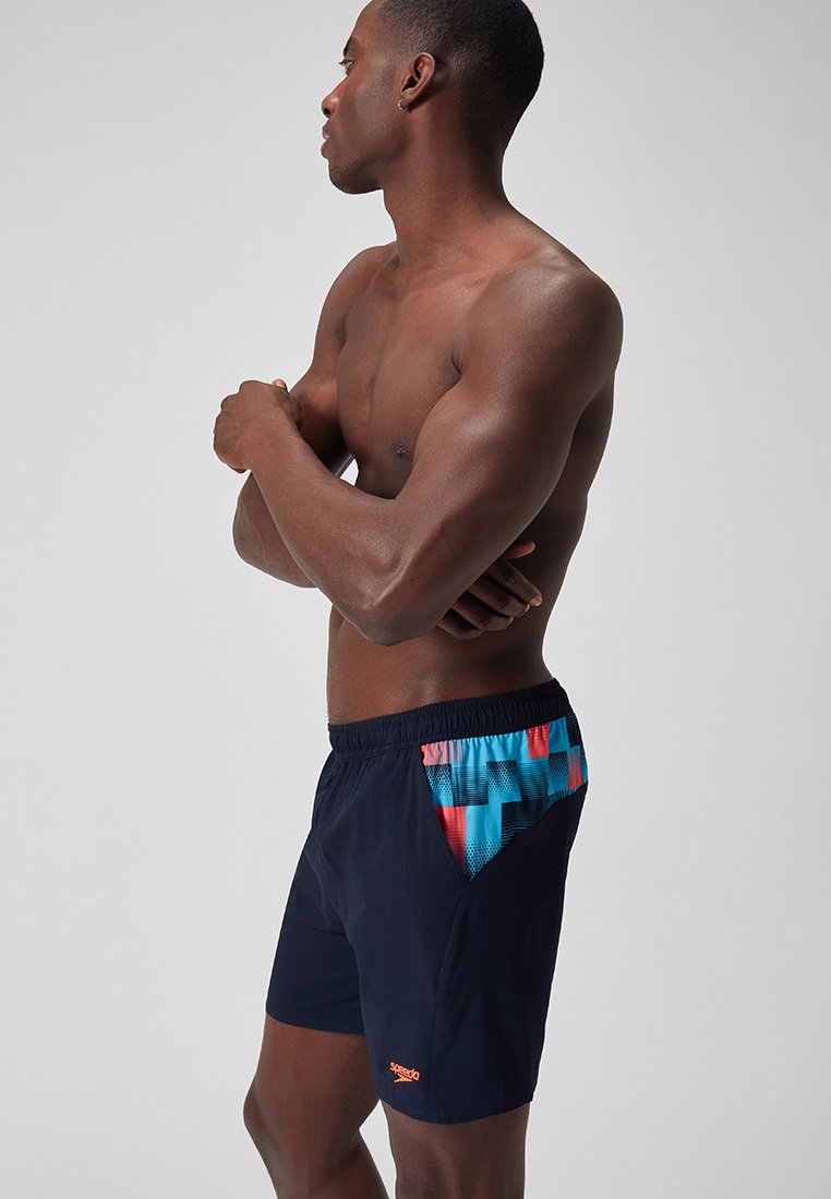 Granatowe kąpielówki z kolorowym geometrycznym wzorem na boku, z elastycznym pasem i pomarańczowym akcentem logo. Gładka faktura.
