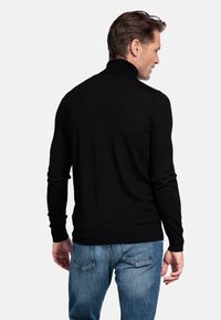 Zwarte gebreide trui met een hoge kraag, getailleerd ontwerp en lange mouwen, gecombineerd met blauwe denimjeans, gezien vanuit de achterkant.