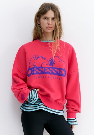 Jeune femme aux longs cheveux portant un sweat-shirt rose avec Snoopy et le texte "OBSESSED GOODNESS & GRIEF" par-dessus une chemise rayée et un pantalon noir.