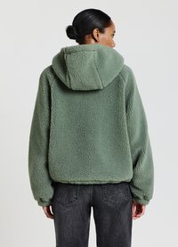 Giacca in pile verde con cappuccio, dotata di una superficie testurizzata, vestibilità ampia e spalle scese, abbinata a jeans grigio scuro.