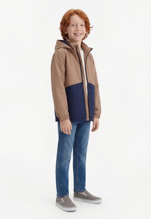 Niño pequeño pelirrojo de pie, con chaqueta con capucha marrón y azul marino, vaqueros azules, camiseta blanca y zapatos grises sin cordones, sonriendo.