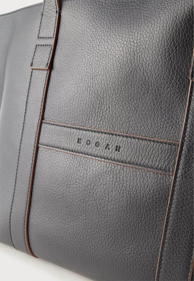 Κοντινή λήψη μαύρης δερμάτινης τσάντας με καφέ ραφές και ανάγλυφο όνομα επωνυμίας "HOGAN" στο εμπρόσθιο πάνελ.