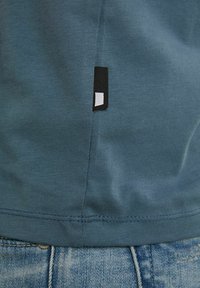 Detailní záběr na lem modro-zelené látkové košile s malou černobílou tkanou etiketou nad pasem modrých džínů.