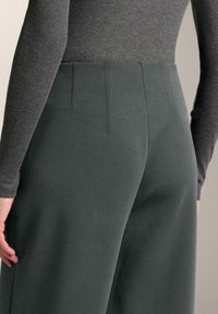 Mörkgröna byxor med vida ben och en mjuk textur, med hög midja och veckdetaljer. En grå ribbad långärmad topp är delvis synlig.