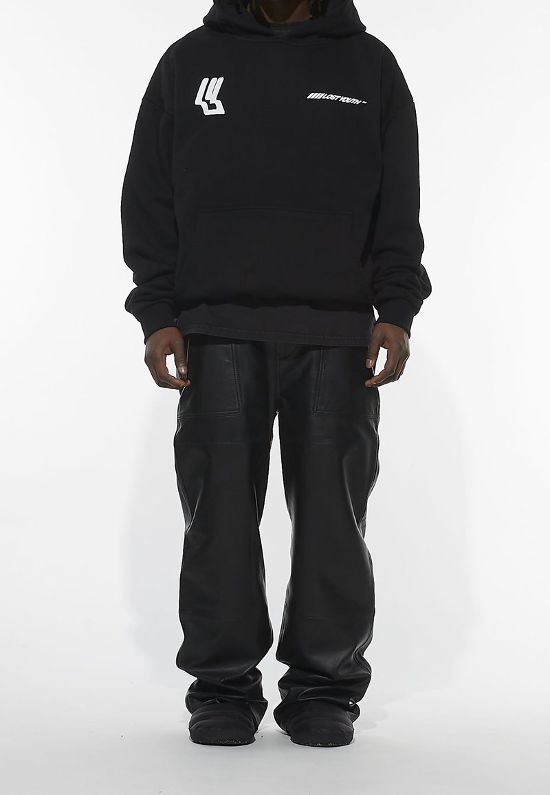 Sweat à capuche noir avec poche avant, orné de logos blancs. Associé à un pantalon en cuir noir ample et des chaussures noires. Fond uni.
