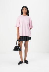 Maglietta di cotone rosa chiaro oversize con maniche corte, abbinata a una gonna nera a plissé, una borsa nera e mocassini neri.