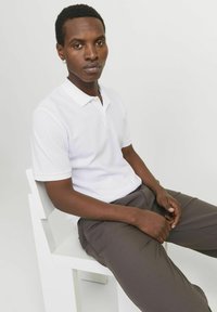 Polo blanc en tissu texturé, avec un col classique et des manches courtes, associé à un pantalon large marron foncé.