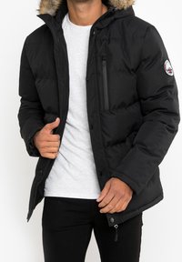 Abrigo puffer negro con capucha con borde de piel, bolsillo en el pecho con cremallera y parche de logo en la manga. Fabricado con un tejido suave y duradero.