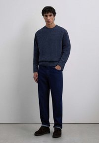 Suéter de punto azul marino con cuello redondo, combinado con jeans de denim oscuro y zapatos de gamuza marrón. Atuendo simple y casual en colores apagados.
