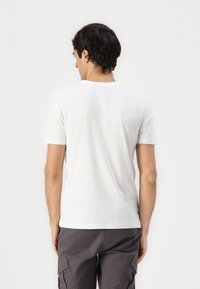 Hvit bomulls t-skjorte med korte ermer og rund hals, enkel design, glatt tekstur, sett fra baksiden. Brukt med grå cargoshorts.