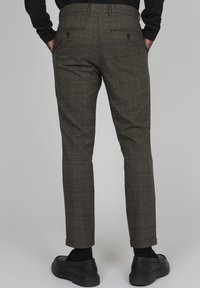 Man med bruna rutiga slim-fit byxor med knäppta bakfickor, svarta strumpor, svarta loafers och en svart långärmad tröja, med händerna i fickorna.