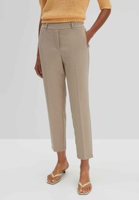 Beige maßgeschneiderte Hose mit slim Fit, frontalen Taschen und einer glatten Textur, kombiniert mit hellen Sandalen. Oben drüber ein gelbes Strickoberteil.