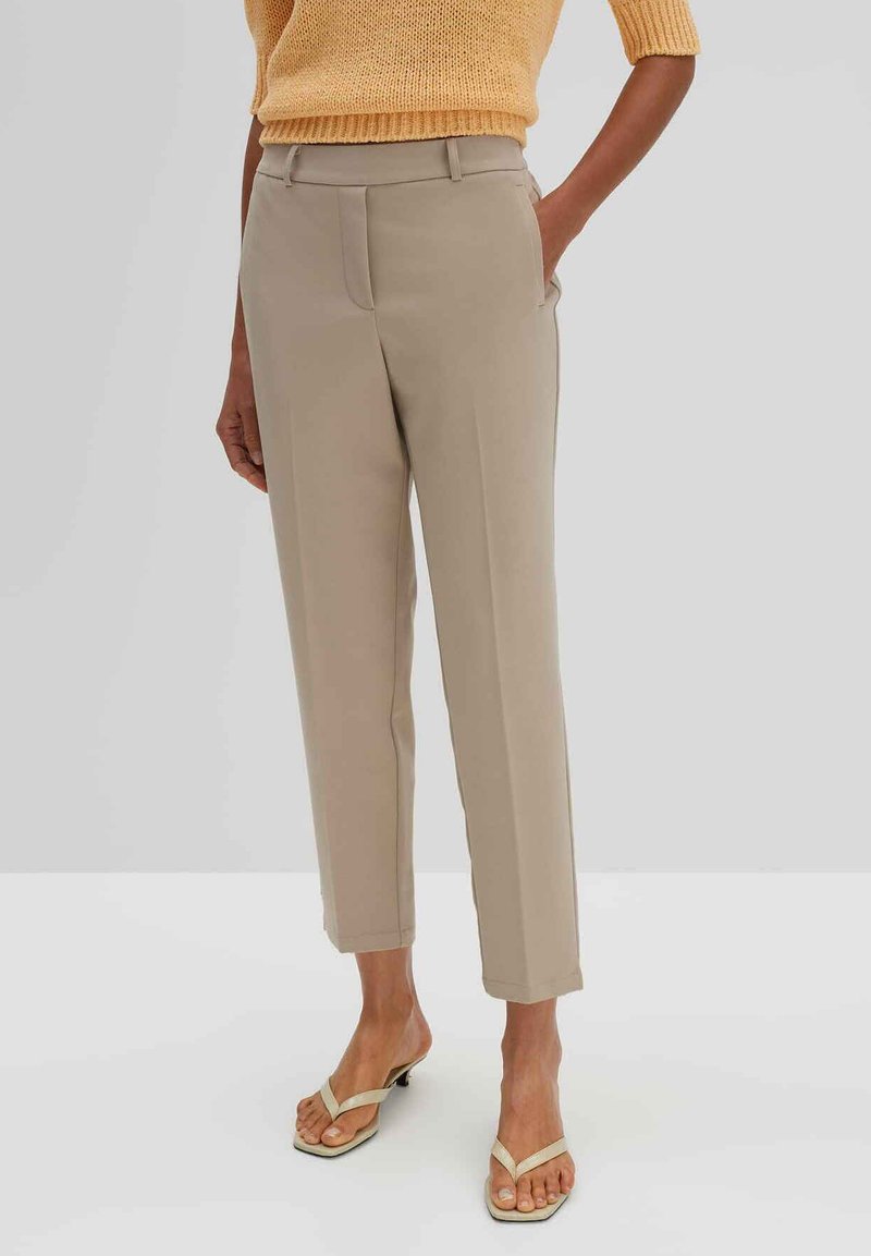 Beige maßgeschneiderte Hose mit slim Fit, frontalen Taschen und einer glatten Textur, kombiniert mit hellen Sandalen. Oben drüber ein gelbes Strickoberteil.