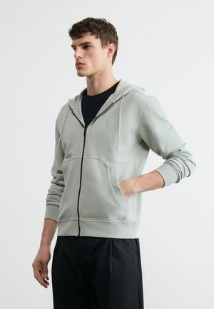 Bărbat tânăr purtând un hanorac deschis la fermoar, gri deschis, cu buzunare frontale și pantaloni negri, stând pe un fundal simplu, deschis la culoare.