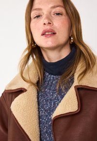 Veste en cuir marron avec col en mouton crème, portée sur un pull bleu texturé. Peau lisse et boucles d'oreilles en or visibles.