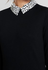 Svart stickad topp med krage, prydd med en vit tygkrage dekorerad med små svarta prickar. Enkel, strukturerad design.