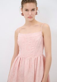 Vestido rosa claro con un corsé ajustado, escote cuadrado y tirantes finos. Presenta pliegues verticales y una falda acampanada con sutiles patrones florales.