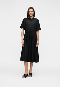 Γυναίκα όρθια, φορώντας μαύρο κουμπωτό φόρεμα μήκους μέχρι το γόνατο με κοντά μανίκια και μαύρα ανοιχτά mules με τακούνι, σε απλό φόντο.