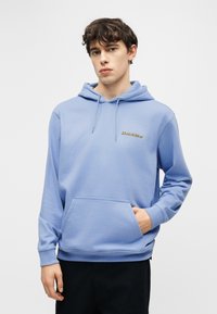 Νεαρός άνδρας που φοράει ένα ανοιχτόγαλανζ Dickies hoodie με μπροστινή τσέπη, στέκεται μπροστά σε ένα απλο λευκό φόντο.