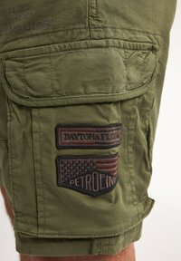 Olive green cargo shorts with two patch pockets, featuring embroidered badges with text and a flag design. Made from durable fabric.