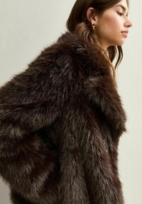 Giovane donna che indossa un cappotto in pelliccia sintetica marrone scuro, in profilo laterale con lunghi capelli castani e orecchini a cerchio dorati su uno sfondo chiaro.
