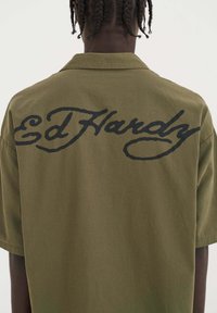 Olijfgroene shirt met korte mouwen met grote zwarte "Ed Hardy" tekst op de rug, gemaakt van lichtgewicht stof met een soepele textuur.