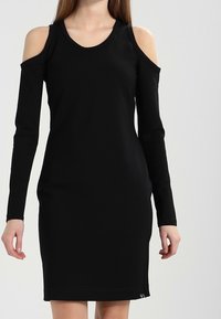 Svart kall axel klänning tillverkad av ett mjukt tyg. Tätt sittande form med långa ärmar, rundad halsringning och subtila nederkant detaljer.