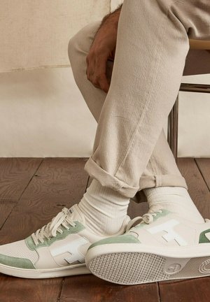 Persoon zittend met gekruiste benen, draagt beige broek, witte sokken en wit-groene sneakers op een houten vloer.