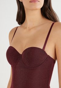 Burgundowy bodysuit z formowanymi miseczkami i regulowanymi ramiączkami, wykonany z błyszczącego, elastycznego materiału. Posiada dekolt w kształcie serca i bezszwowe krawędzie.