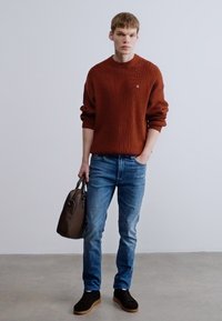 Barna bordázott pulóver, kék farmer, fekete tornacipő gumitalppal és barna táska. A ruházat laza szabású és minimalista részleteket tartalmaz.