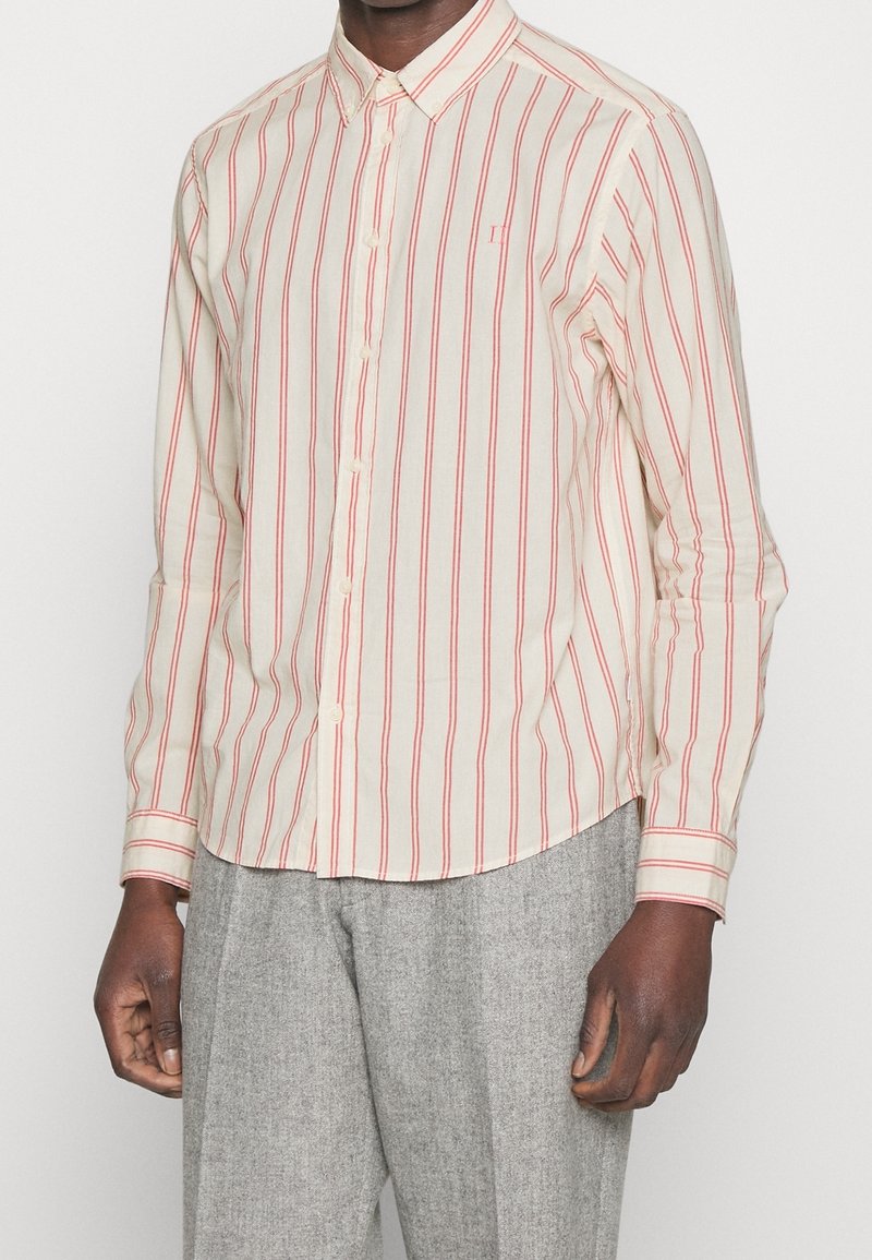 Camisa de manga larga en color crema con rayas verticales rojas, cuello abotonado y dobladillo redondeado. Presenta un pequeño logo en el pecho. La tela parece ligera.