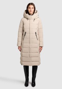 Béžový prešívaný kabát s kapucňou, so zipsom vpredu, bočnými vreckami a horizontálnym prešívaným vzorom. Hladká textúra. Nosí sa cez čierne nohavice.
